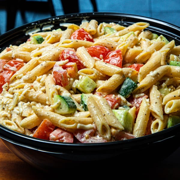 Close-up of a pasta salad with penne, tomatoes, cucumbers, and creamy dressing in a black bowl.